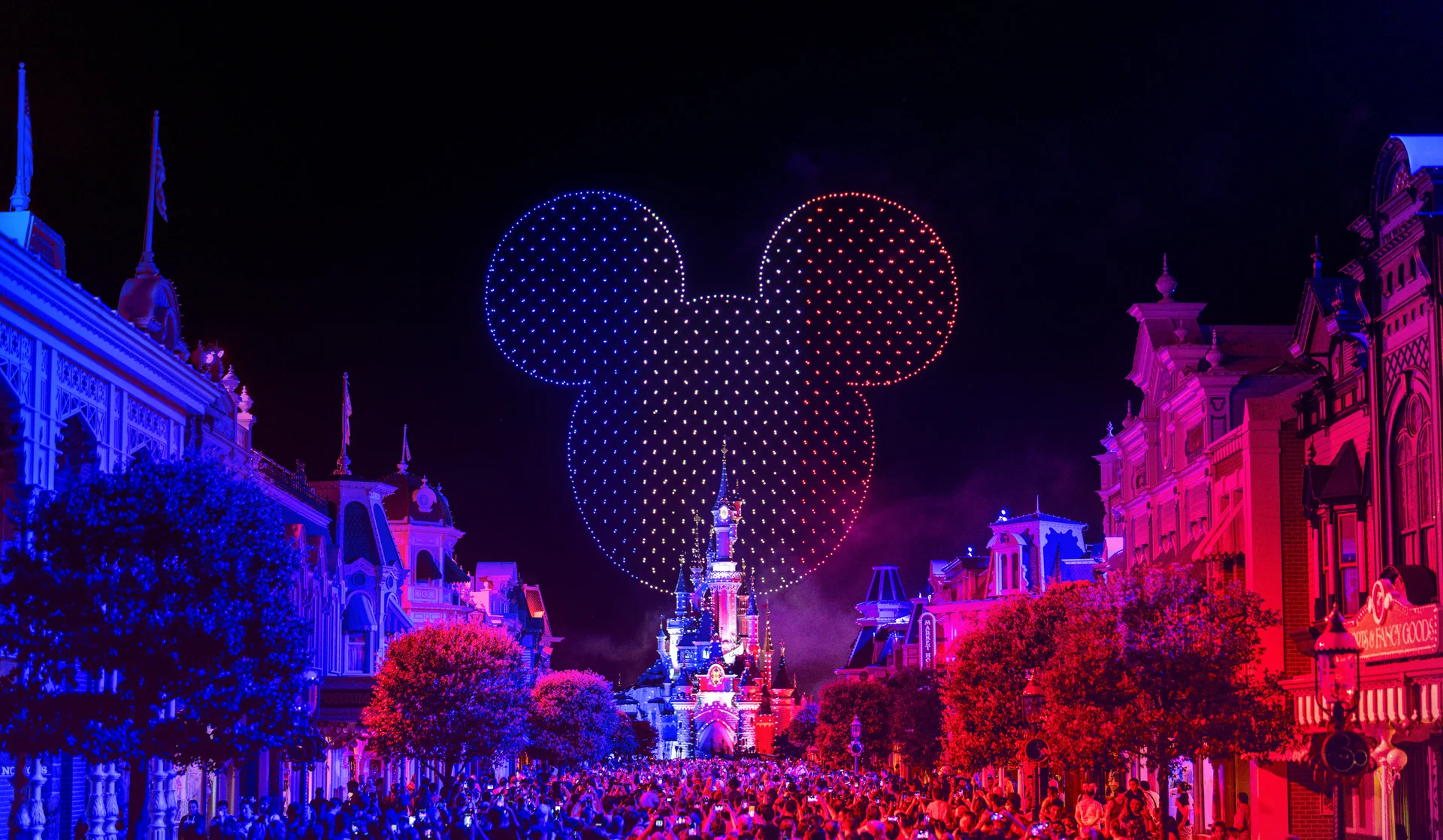 Un « Spectacle Nocturne » éblouissant à Disneyland Paris projette une immense silhouette représentant la tête de Mickey Mouse au-dessus du château, tandis que les bâtiments sont illuminés de bleu et de rouge et qu'une foule s'est rassemblée en contrebas pour célébrer le 14 juillet.