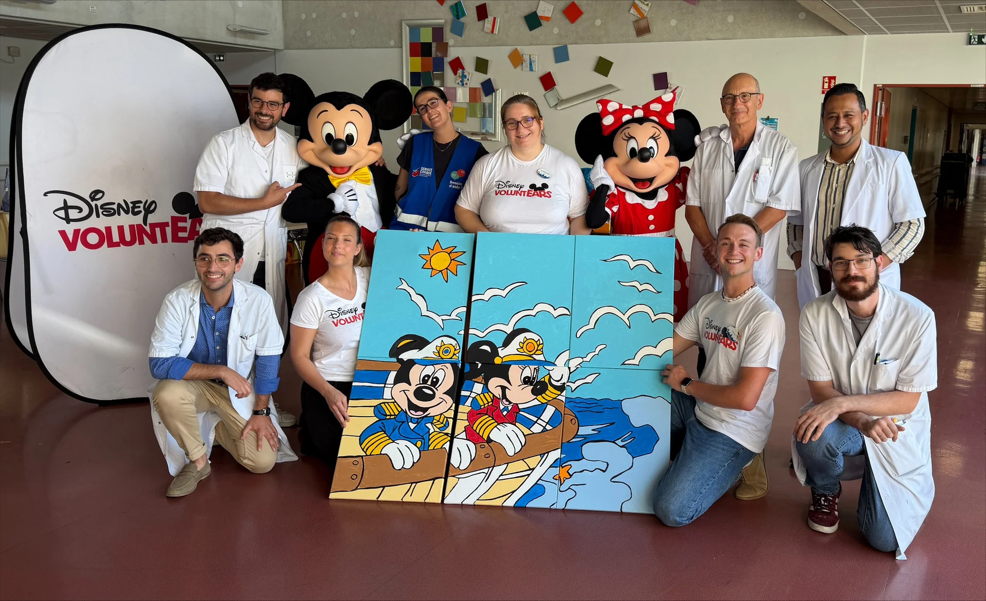 Un groupe de personnes, certaines portant des t-shirts de bénévoles Disney et d'autres des blouses de laboratoire, posent aux côtés des mascottes Mickey et Minnie Mouse ainsi que d'un grand puzzle représentant Mickey et Minnie sur un bateau, pour célébrer une collaboration exceptionnelle entre Disneyland Paris et le CHU de Toulouse.