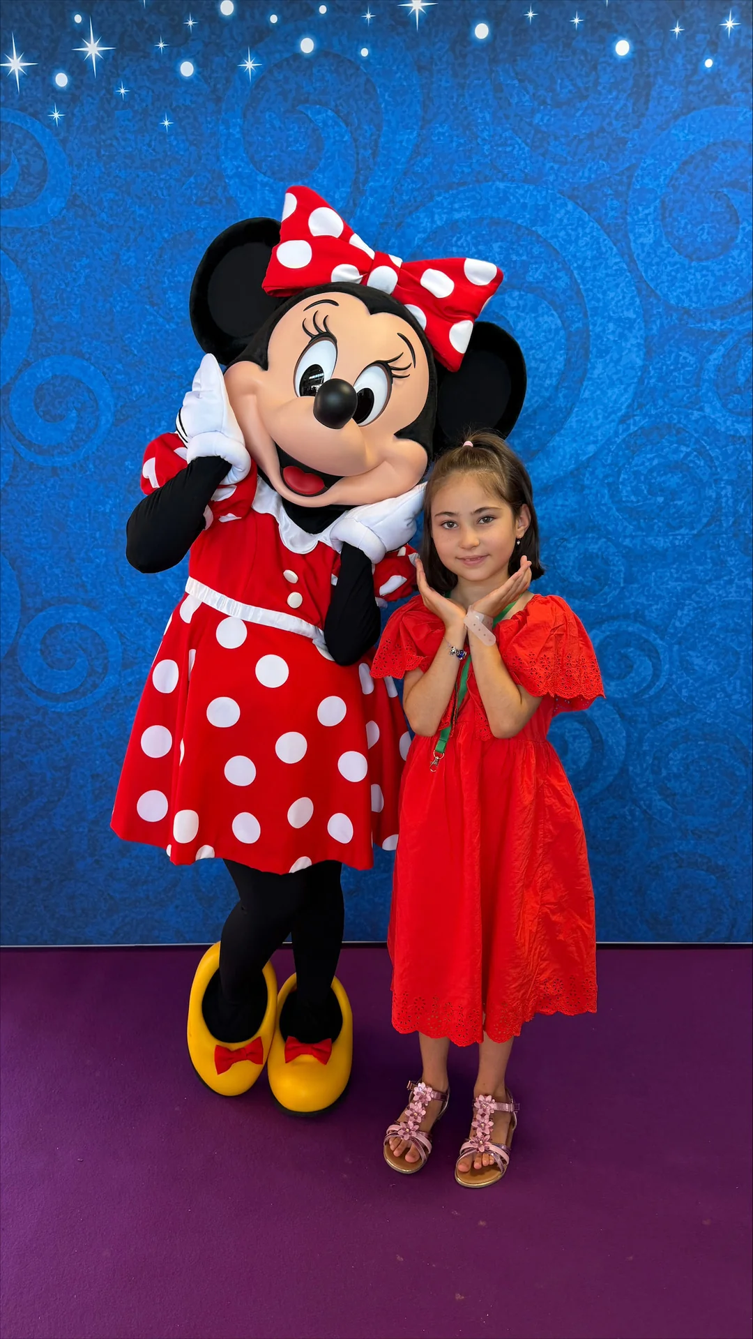 Une petite fille en robe rouge pose aux côtés d'une personne déguisée en Minnie Mouse, devant un fond à motifs bleus, capturant ainsi la magie de Mickey et Minnie, comme s'ils se trouvaient à Disneyland Paris.