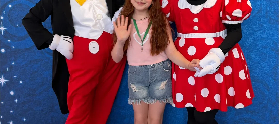 Une jeune fille se tient au milieu d'un groupe de personnes déguisées en Mickey Mouse et Minnie Mouse ; elle sourit et fait signe de la main, devant un fond bleu parsemé d'étoiles, lors d'un événement organisé par le CHU de Toulouse et inspiré par la magie de Disneyland Paris.