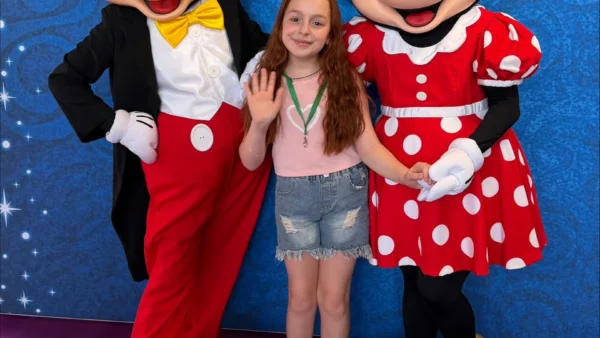 Une jeune fille se tient au milieu d'un groupe de personnes déguisées en Mickey Mouse et Minnie Mouse ; elle sourit et fait signe de la main, devant un fond bleu parsemé d'étoiles, lors d'un événement organisé par le CHU de Toulouse et inspiré par la magie de Disneyland Paris.