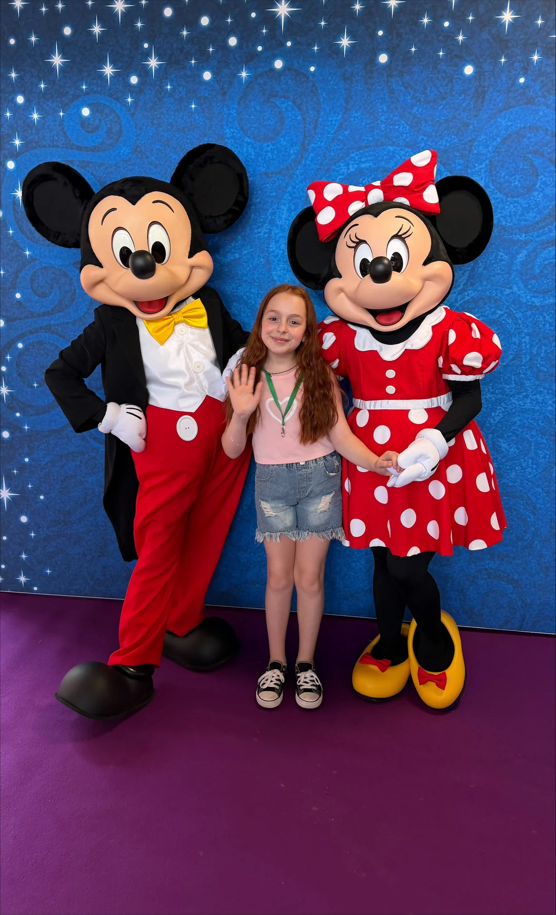 Une jeune fille se tient au milieu d'un groupe de personnes déguisées en Mickey Mouse et Minnie Mouse ; elle sourit et fait signe de la main, devant un fond bleu parsemé d'étoiles, lors d'un événement organisé par le CHU de Toulouse et inspiré par la magie de Disneyland Paris.