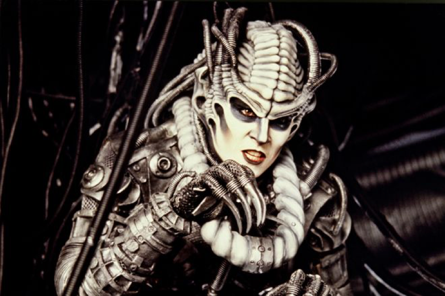Une personne vêtue d'un costume de science-fiction sophistiqué inspiré du Capitaine EO, avec un maquillage pâle, une armure mécanique, des câbles et des gants en forme de griffes, sur un fond sombre parsemé d'une multitude de câbles entrelacés — rappelant le style avant-gardiste en vogue jusqu'à sa fermeture, le 17 août 1998.