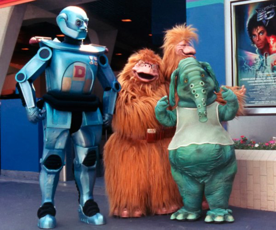 Quatre personnes vêtues de costumes colorés — un robot argenté, deux créatures à fourrure et un personnage vert ressemblant à un éléphant — se tiennent côte à côte devant un bâtiment, avec une affiche de film en arrière-plan, ce qui rappelle l'époque précédant la fermeture de Captain EO, le 17 août 1998.