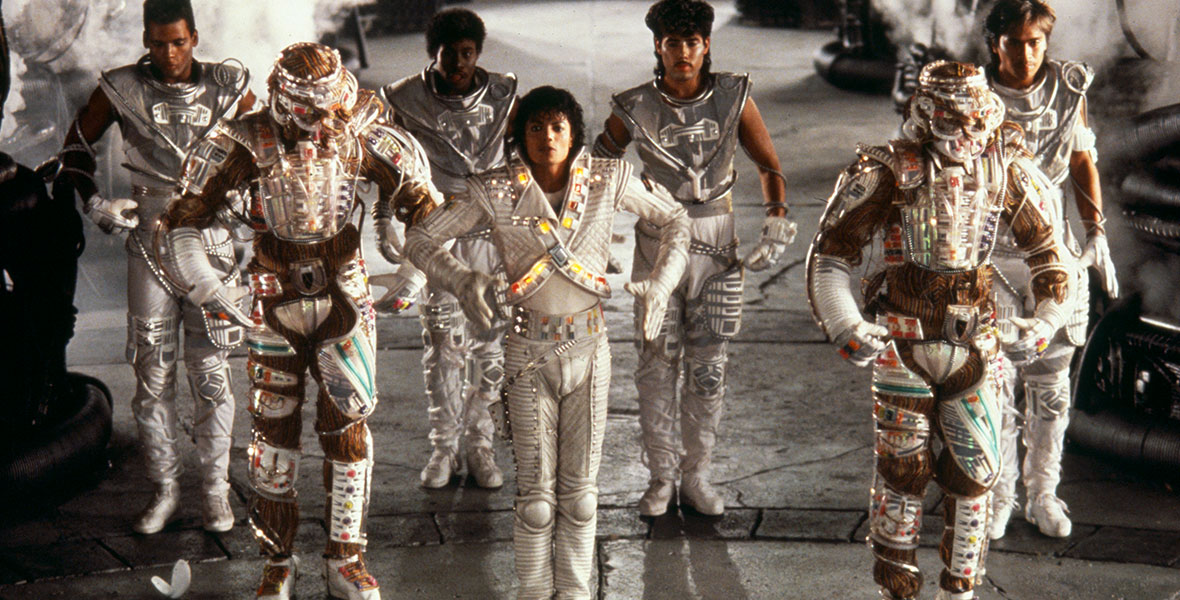 Six personnes vêtues de costumes métalliques futuristes se tiennent en formation ; deux portent des armures sophistiquées, quatre des tenues argentées épurées, évoquant l’esprit de science-fiction de Captain EO sur un fond brumeux et dans un décor rappelant une fin d’un autre monde — peut-être un clin d’œil à la période autour du 17 août 1998.