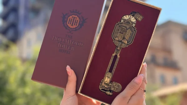 Deux mains tiennent une boîte décorative représentant l'Hollywood Tower Hotel et une clé dorée ouvragée devant un bâtiment en plein air, capturant ainsi l'atmosphère emblématique de la Twilight Zone Tower of Terror à Disneyland Paris. Cet objet de collection en édition limitée est un incontournable pour les fans à la recherche d'une touche de magie Disney et d'une élégance mystérieuse.