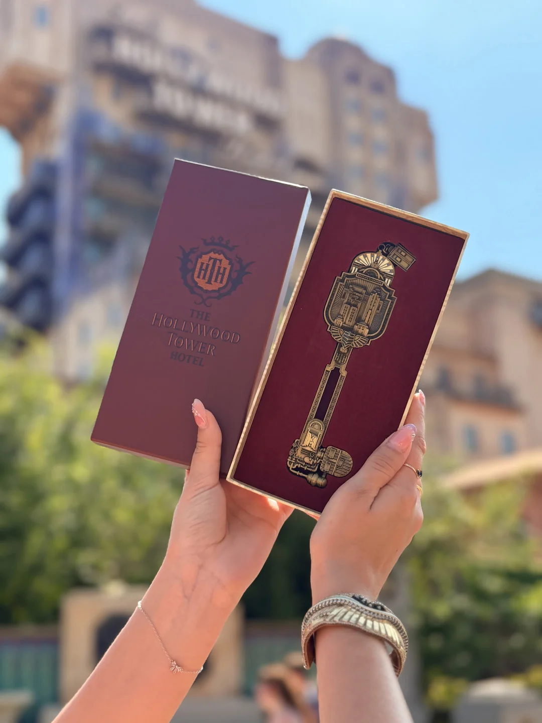 Deux mains tiennent une boîte décorative représentant l'Hollywood Tower Hotel et une clé dorée ouvragée devant un bâtiment en plein air, capturant ainsi l'atmosphère emblématique de la Twilight Zone Tower of Terror à Disneyland Paris. Cet objet de collection en édition limitée est un incontournable pour les fans à la recherche d'une touche de magie Disney et d'une élégance mystérieuse.