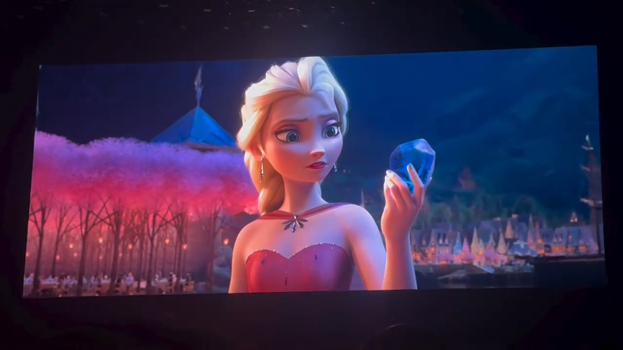 Un personnage d'animation aux cheveux blonds, vêtu d'une robe rouge, tient un grand cristal bleu, avec en arrière-plan des arbres roses et un paysage urbain nocturne — une scène qui rappelle les moments magiques de Disney dévoilés lors du D23 et qui inspirent de futurs projets.