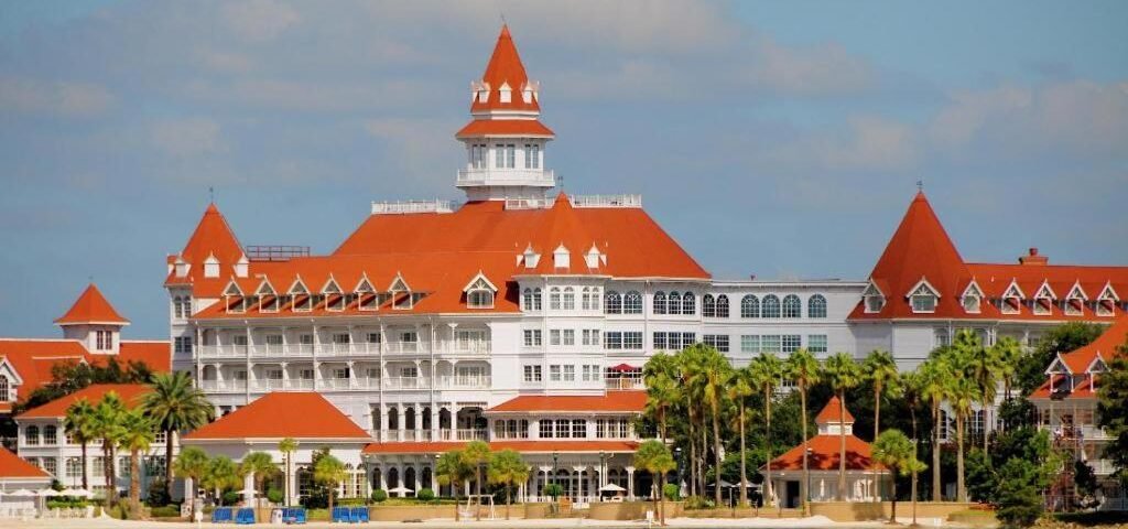 Grand complexe hôtelier blanc de style victorien, aux toits rouges, doté de nombreux balcons et de tours, situé au bord d'un front de mer bordé de palmiers sous un ciel partiellement nuageux — souvent considéré comme le plus bel hôtel Disney et fréquemment salué comme le meilleur hôtel Disney pour son élégance et son emplacement exceptionnel.