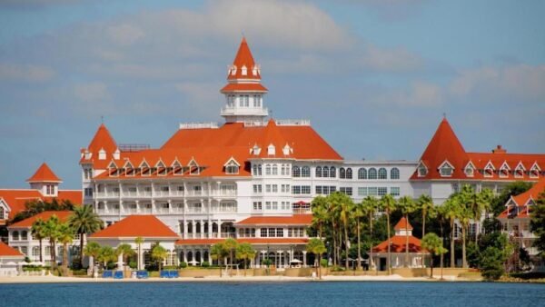 Grand complexe hôtelier blanc de style victorien, aux toits rouges, doté de nombreux balcons et de tours, situé au bord d'un front de mer bordé de palmiers sous un ciel partiellement nuageux — souvent considéré comme le plus bel hôtel Disney et fréquemment salué comme le meilleur hôtel Disney pour son élégance et son emplacement exceptionnel.