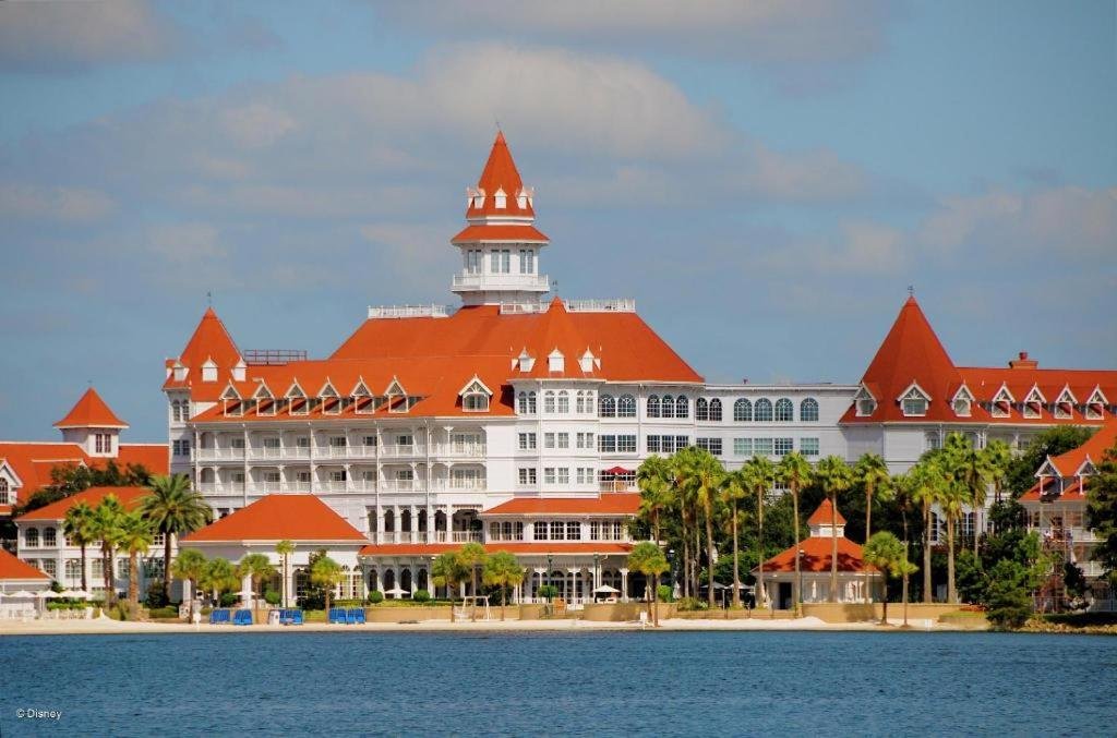 Grand complexe hôtelier blanc de style victorien, aux toits rouges, doté de nombreux balcons et de tours, situé au bord d'un front de mer bordé de palmiers sous un ciel partiellement nuageux — souvent considéré comme le plus bel hôtel Disney et fréquemment salué comme le meilleur hôtel Disney pour son élégance et son emplacement exceptionnel.