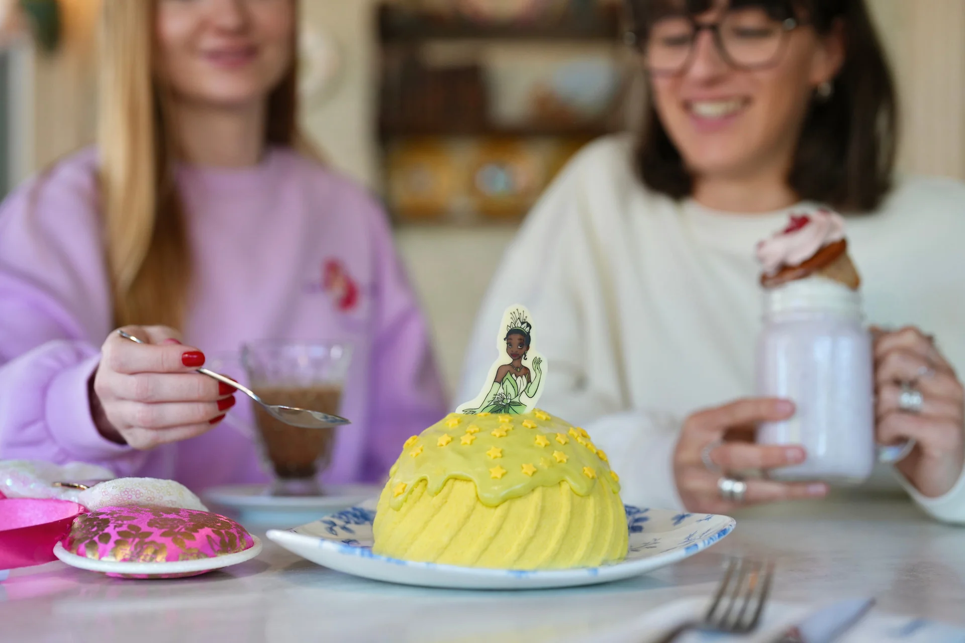 Deux femmes sont assises à une table devant des boissons et un gâteau en forme de dôme jaune orné d'une décoration représentant la princesse Tiana, pour célébrer la Semaine internationale des princesses. L'une tient une fourchette, tandis que l'autre savoure un milkshake garni de crème fouettée et de garnitures, illustrant ainsi l'atmosphère enchanteresse de Disneyland Paris en 2026.