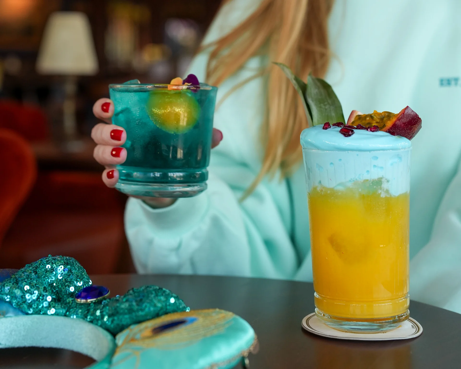 Sur une table, une personne tient un cocktail bleu avec des glaçons et une garniture jaune, à côté d'un cocktail jaune recouvert d'une mousse bleue et agrémenté de fruits, illustrant ainsi l'esprit magique de Disneyland Paris lors de la merveilleuse Semaine internationale des princesses de 2026.