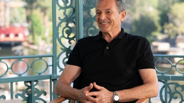Un homme souriant, vêtu d'un polo noir, est assis sur une chaise à l'extérieur, avec en arrière-plan des ferronneries bleu sarcelle ouvragées et des arbres flous — une image qui incarne l'esprit décontracté des « Worldbuilders » et des « créateurs » de Disney.