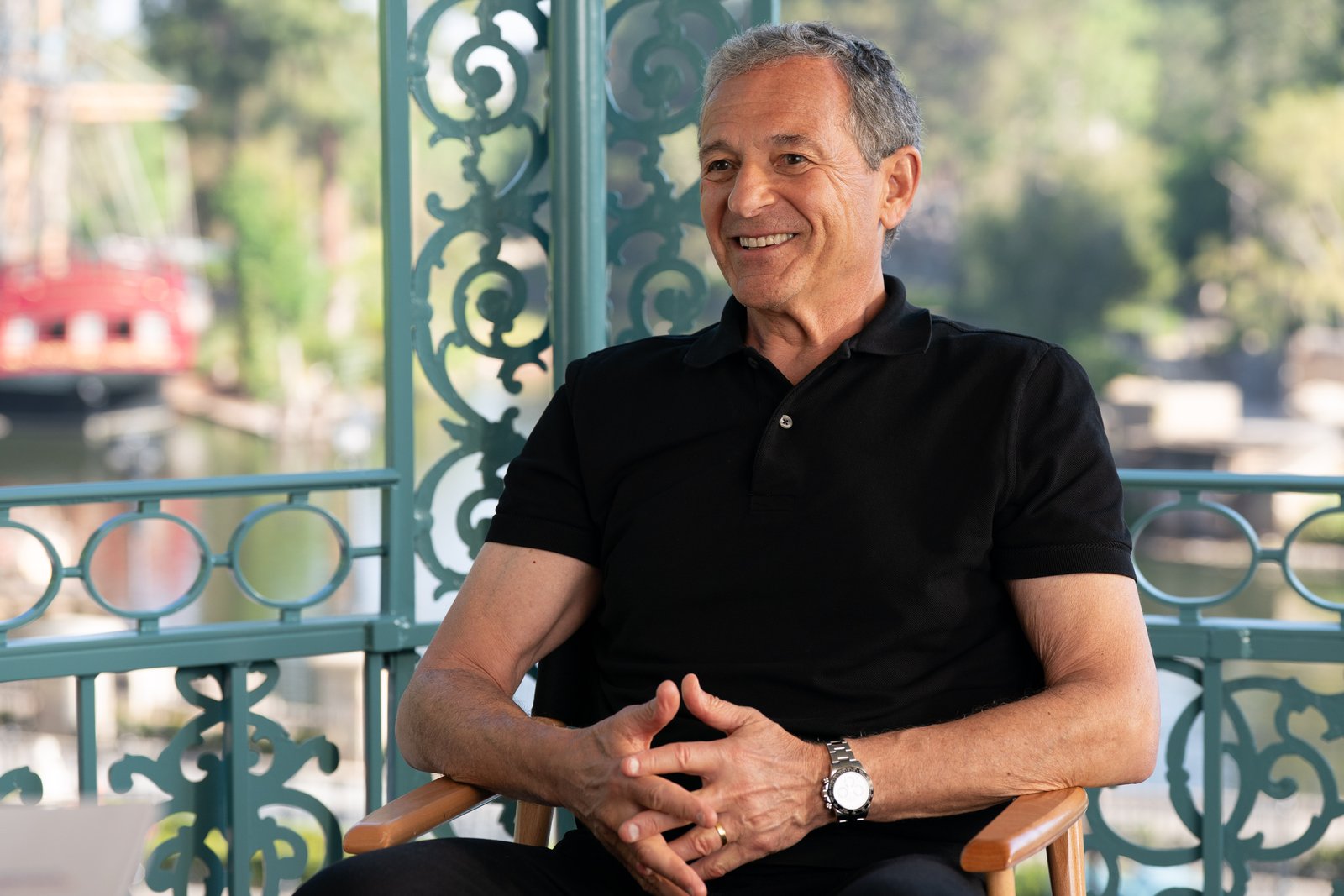 Un homme souriant, vêtu d'un polo noir, est assis sur une chaise à l'extérieur, avec en arrière-plan des ferronneries bleu sarcelle ouvragées et des arbres flous — une image qui incarne l'esprit décontracté des « Worldbuilders » et des « créateurs » de Disney.