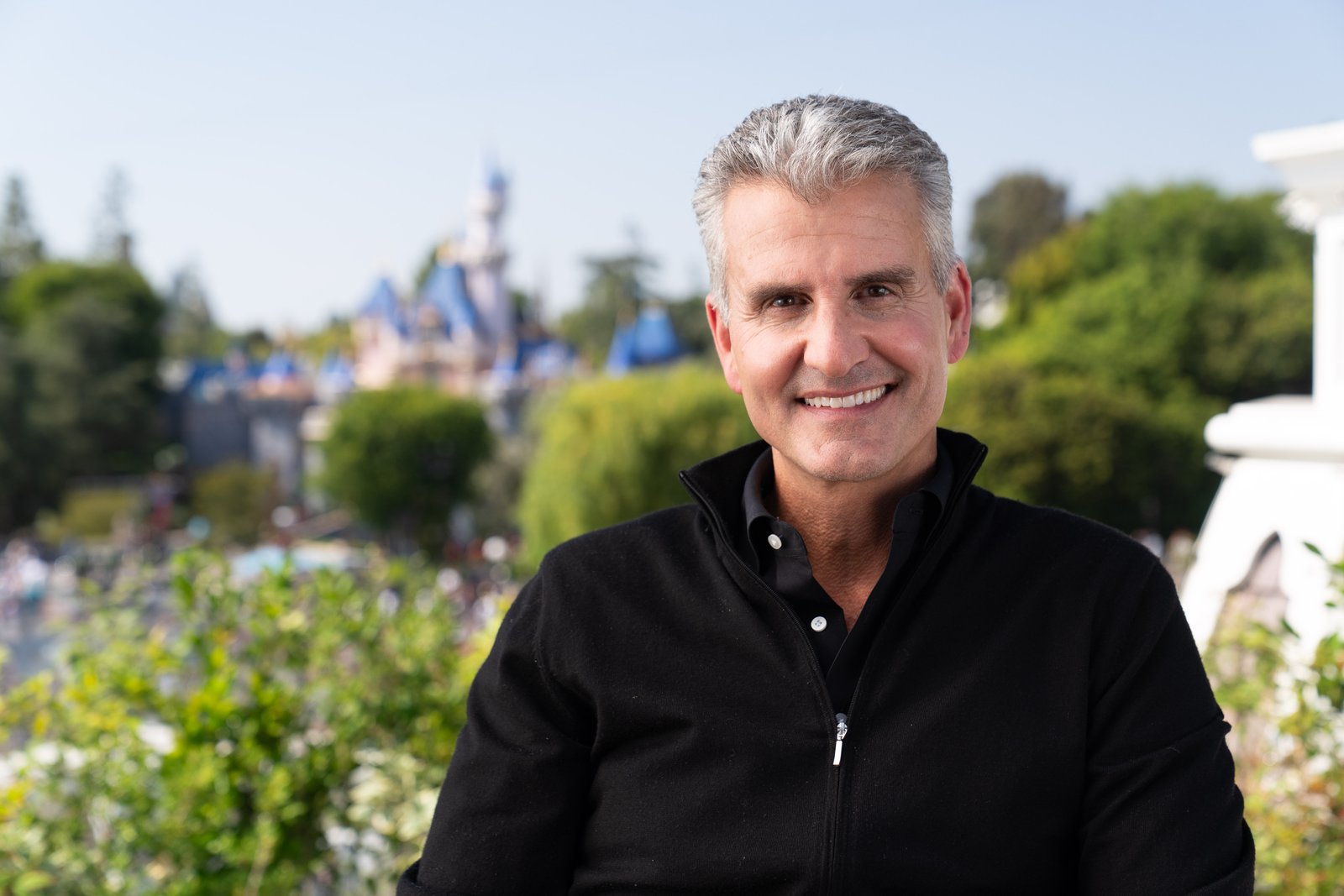 Un homme aux cheveux gris, vêtu d'un pull noir à fermeture éclair, sourit, assis en plein air, avec en arrière-plan un château flou et de la verdure — une image qui incarne l'esprit des « Worldbuilders » de Disney et des « créateurs de la magie » Disney.