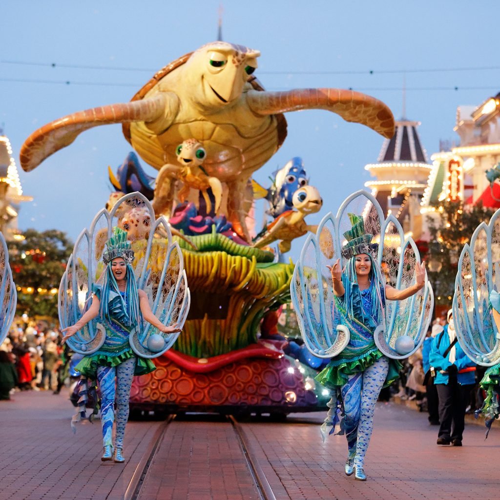 Des artistes vêtus de costumes bleus dansent devant un immense char de parade représentant des personnages du film « Le Monde de Nemo », dont une tortue de mer géante, lors de la nouvelle parade organisée à Disneyland Paris, pour le plus grand plaisir des visiteurs dans une rue du parc d'attractions en 2028.
