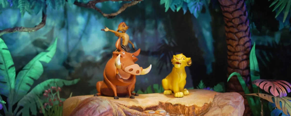 Trois personnages d'animaux de dessin animé sont assis sur une bûche dans un décor de jungle : un suricate se tient debout sur un phacochère, tandis qu'un lion est assis à proximité. Inspirée des maquettes du Roi Lion, cette scène colorée et éclairée par des projecteurs de scène évoque la magie d'un « futur monde immersif ».