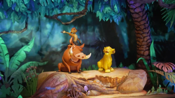 Trois personnages d'animaux de dessin animé sont assis sur une bûche dans un décor de jungle : un suricate se tient debout sur un phacochère, tandis qu'un lion est assis à proximité. Inspirée des maquettes du Roi Lion, cette scène colorée et éclairée par des projecteurs de scène évoque la magie d'un « futur monde immersif ».