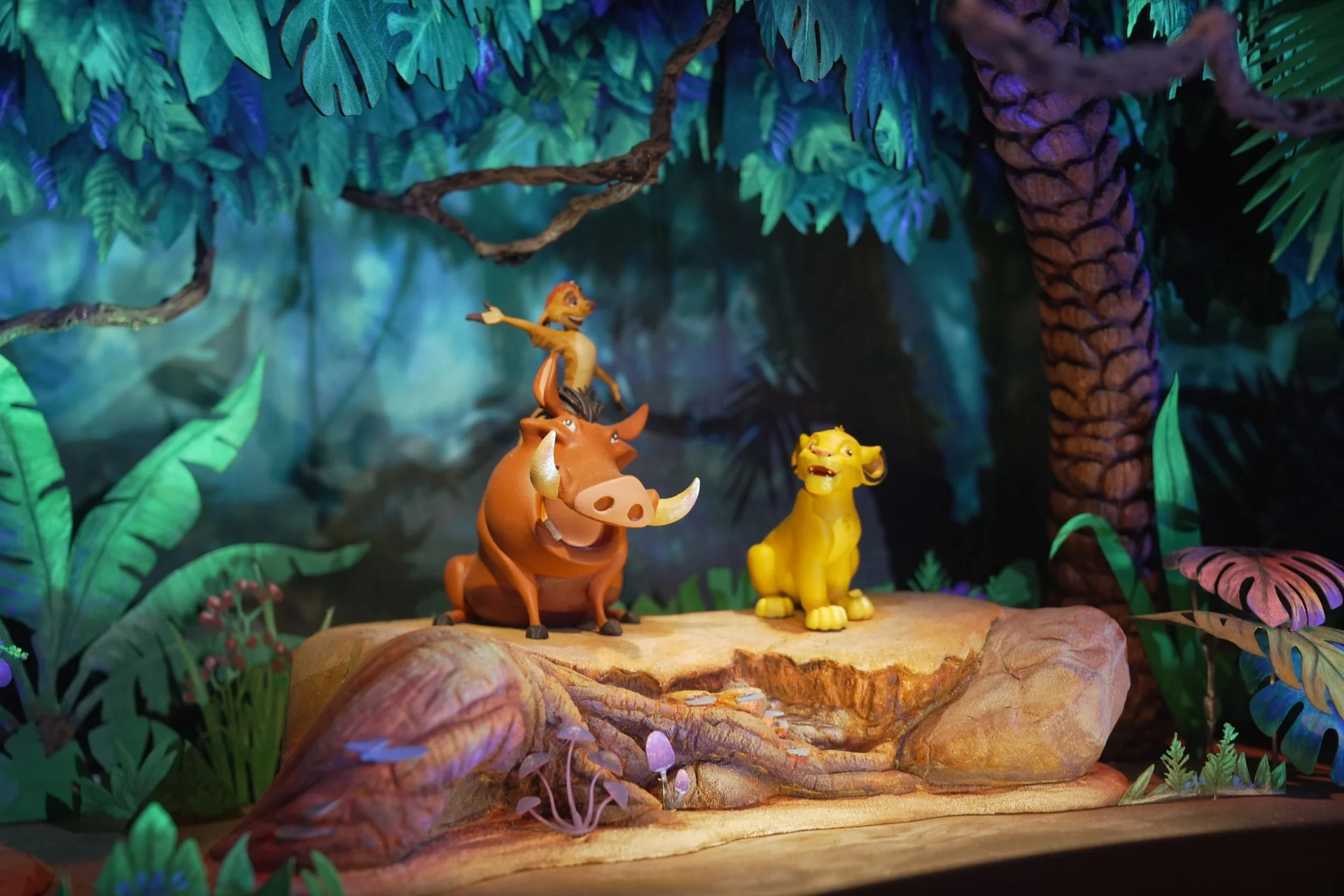 Trois personnages d'animaux de dessin animé sont assis sur une bûche dans un décor de jungle : un suricate se tient debout sur un phacochère, tandis qu'un lion est assis à proximité. Inspirée des maquettes du Roi Lion, cette scène colorée et éclairée par des projecteurs de scène évoque la magie d'un « futur monde immersif ».