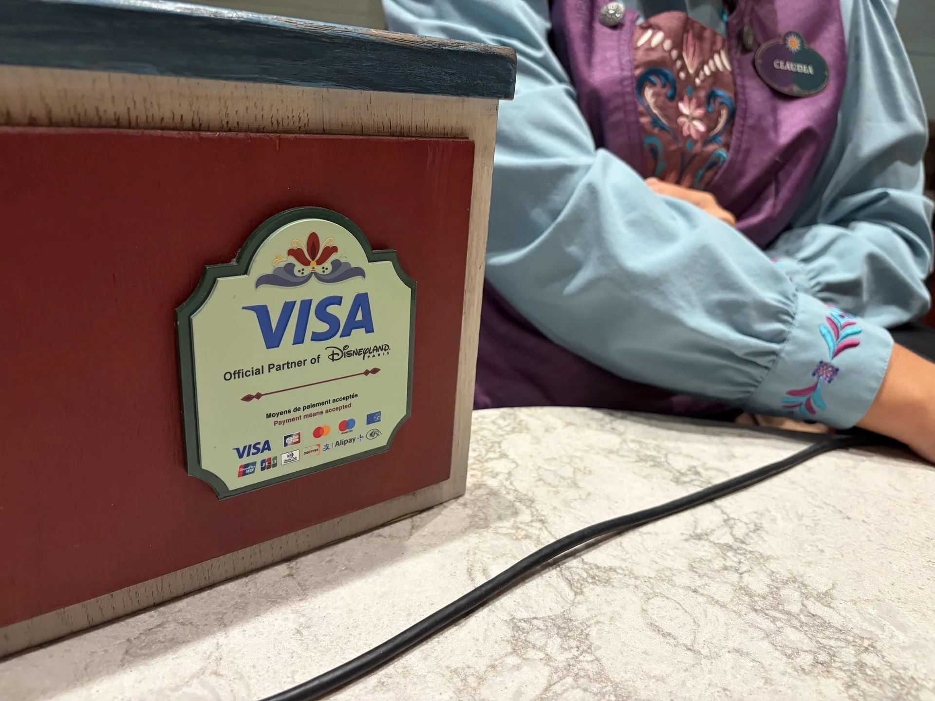 Une personne vêtue d'un uniforme bleu et violet se tient à côté d'un terminal de paiement rouge, sur lequel figurent le logo Visa et les types de cartes acceptées, posé sur un comptoir en marbre à Disneyland Paris.