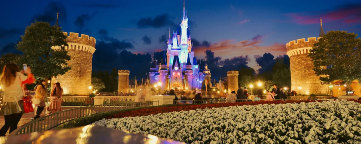 La nuit, à Disneyland Tokyo, au Japon, des visiteurs se promènent près d’un château brillamment illuminé, entouré de tours, de jardins parsemés de fleurs blanches, avec en arrière-plan un ciel bleu foncé parsemé de nuages colorés — créant ainsi une atmosphère pleine de charme et d’enchantement.