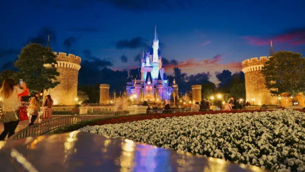 La nuit, à Disneyland Tokyo, au Japon, des visiteurs se promènent près d’un château brillamment illuminé, entouré de tours, de jardins parsemés de fleurs blanches, avec en arrière-plan un ciel bleu foncé parsemé de nuages colorés — créant ainsi une atmosphère pleine de charme et d’enchantement.