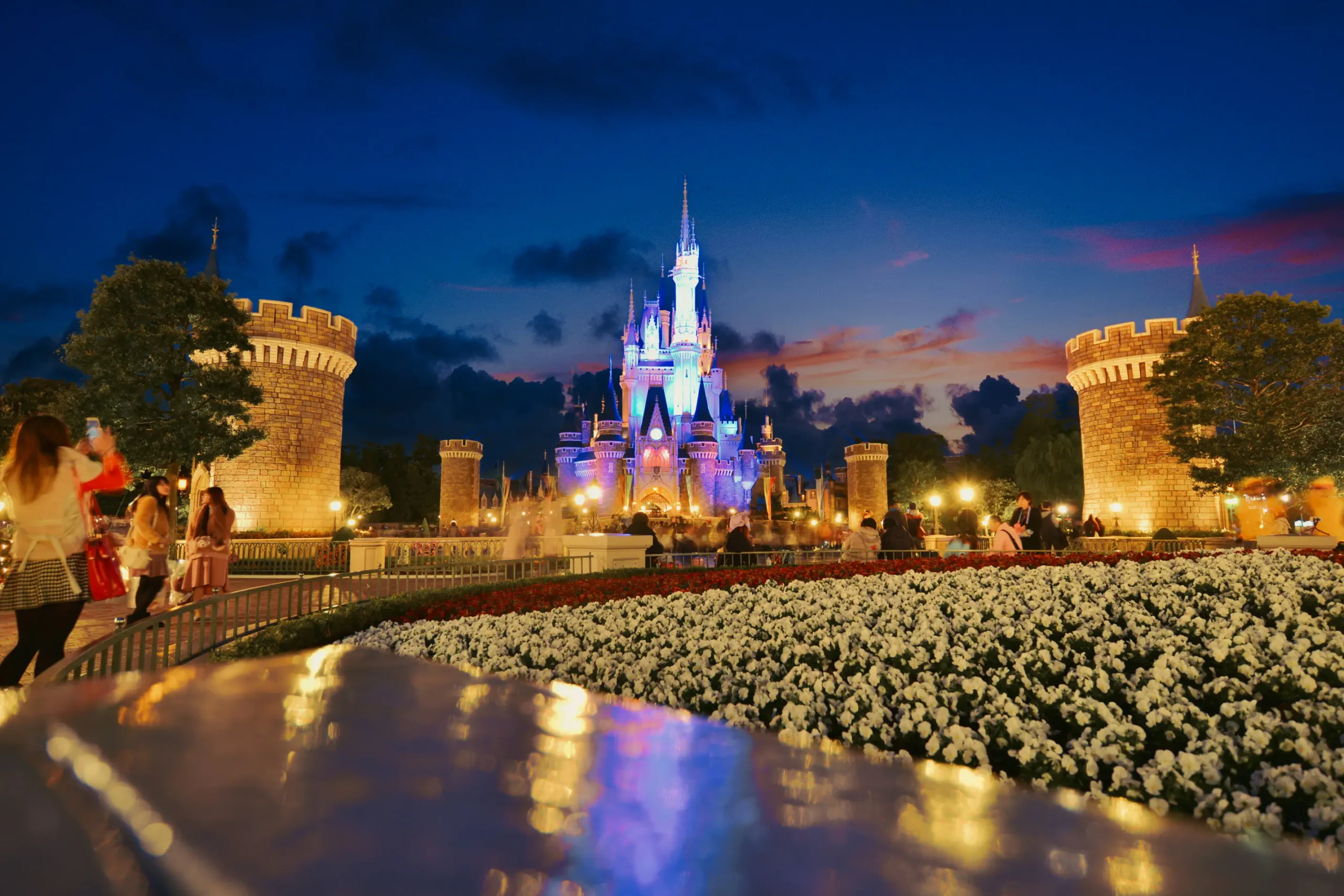La nuit, à Disneyland Tokyo, au Japon, des visiteurs se promènent près d’un château brillamment illuminé, entouré de tours, de jardins parsemés de fleurs blanches, avec en arrière-plan un ciel bleu foncé parsemé de nuages colorés — créant ainsi une atmosphère pleine de charme et d’enchantement.