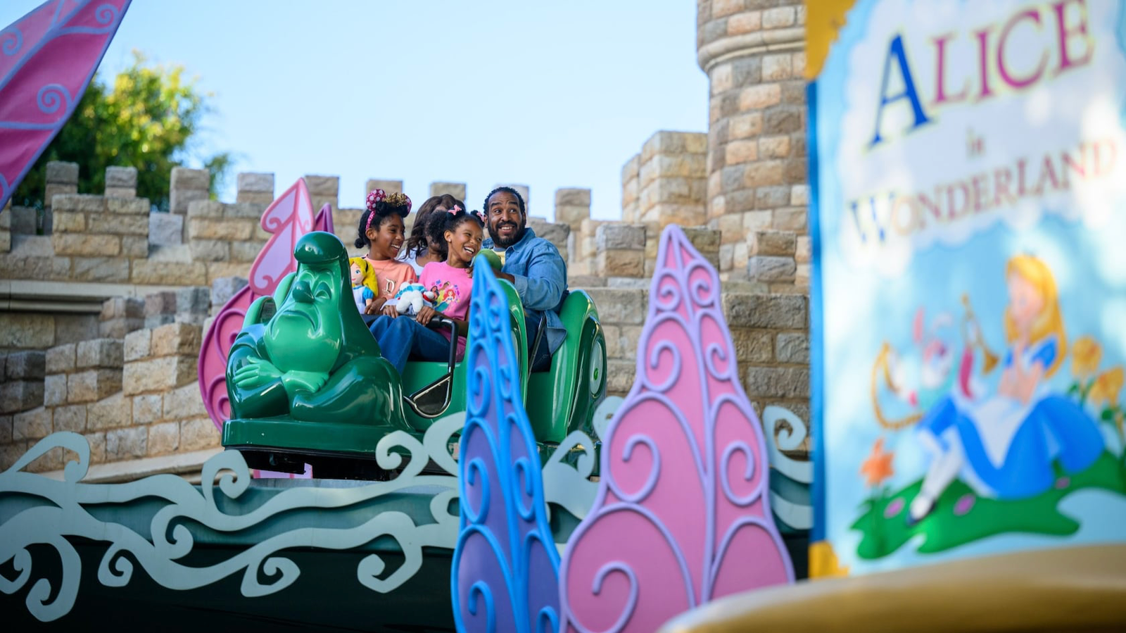 Un homme et deux enfants montent à bord d'une voiturette verte en forme de chenille sur l'attraction « Alice au pays des merveilles » d'un parc d'attractions ; au premier plan, on aperçoit le panneau de l'attraction, qui indique son inauguration le 14 juin 1958.