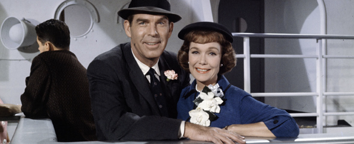 Un homme en costume et chapeau et une femme vêtue d'une tenue bleue ornée de fleurs blanches se tiennent côte à côte sur le pont d'un bateau, souriant à la caméra comme s'ils s'apprêtaient à dire « Bon voyage » avant de se lancer dans leur périple — une image qui rappelle un film classique du 17 mai 1962.