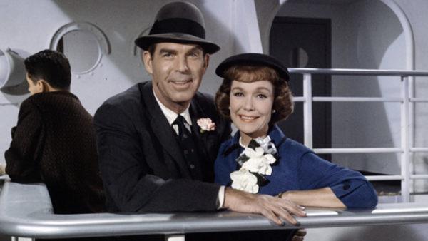 Un homme en costume et chapeau et une femme vêtue d'une tenue bleue ornée de fleurs blanches se tiennent côte à côte sur le pont d'un bateau, souriant à la caméra comme s'ils s'apprêtaient à dire « Bon voyage » avant de se lancer dans leur périple — une image qui rappelle un film classique du 17 mai 1962.