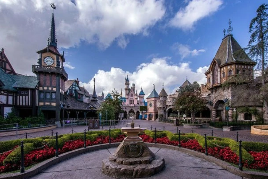Vue d'un château et des bâtiments environnants dans le New Fantasyland de Disneyland, avec au premier plan un parterre de fleurs circulaire et un cadran solaire sous un ciel partiellement nuageux, capturant l'atmosphère enchanteresse telle qu'elle aurait pu se présenter le 25 mai 1983.