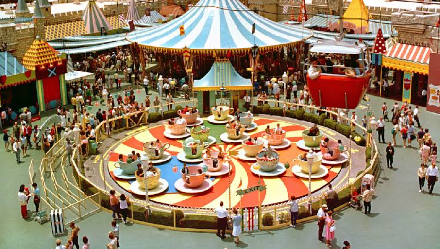 Dans un parc d'attractions, un manège coloré en forme de tasses tournantes est pris d'assaut par la foule, tandis qu'on aperçoit des tentes et des stands en arrière-plan : une scène animée qui rappelle l'inauguration du New Fantasyland à Disneyland, le 25 mai 1983.