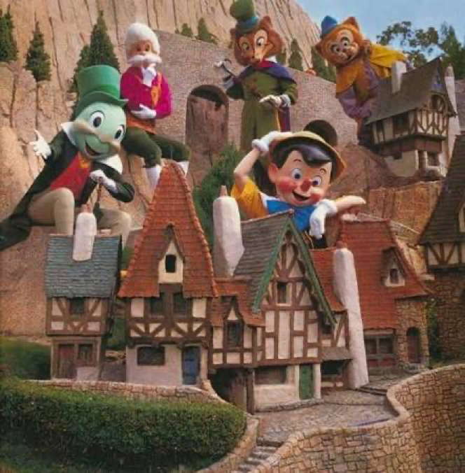 Cinq personnages costumés, dont Pinocchio et Jiminy Cricket, posent au sommet et autour d'une maquette détaillée représentant un village composé de maisons de style cottage et agrémenté de verdure, incarnant ainsi l'atmosphère enchanteresse de New Fantasyland lors de son inauguration à Disneyland.