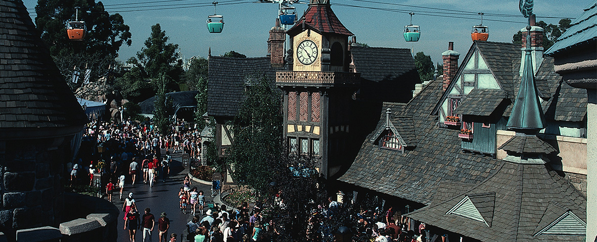Une foule se promène dans un village à thème situé dans le New Fantasyland de Disneyland, où l’on peut admirer des gondoles colorées suspendues dans les airs et une tour horloge au centre. L’ambiance y est animée et trépidante, reflétant toute la magie qui attire les visiteurs depuis le 25 mai 1983, tandis que les rues débordantes de vie grouillent de monde.