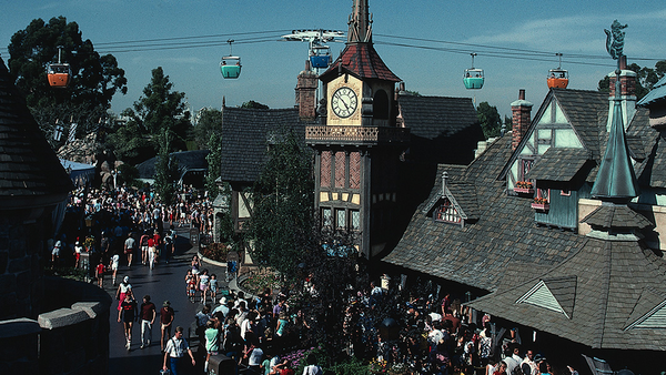 Une foule se promène dans un village à thème situé dans le New Fantasyland de Disneyland, où l’on peut admirer des gondoles colorées suspendues dans les airs et une tour horloge au centre. L’ambiance y est animée et trépidante, reflétant toute la magie qui attire les visiteurs depuis le 25 mai 1983, tandis que les rues débordantes de vie grouillent de monde.