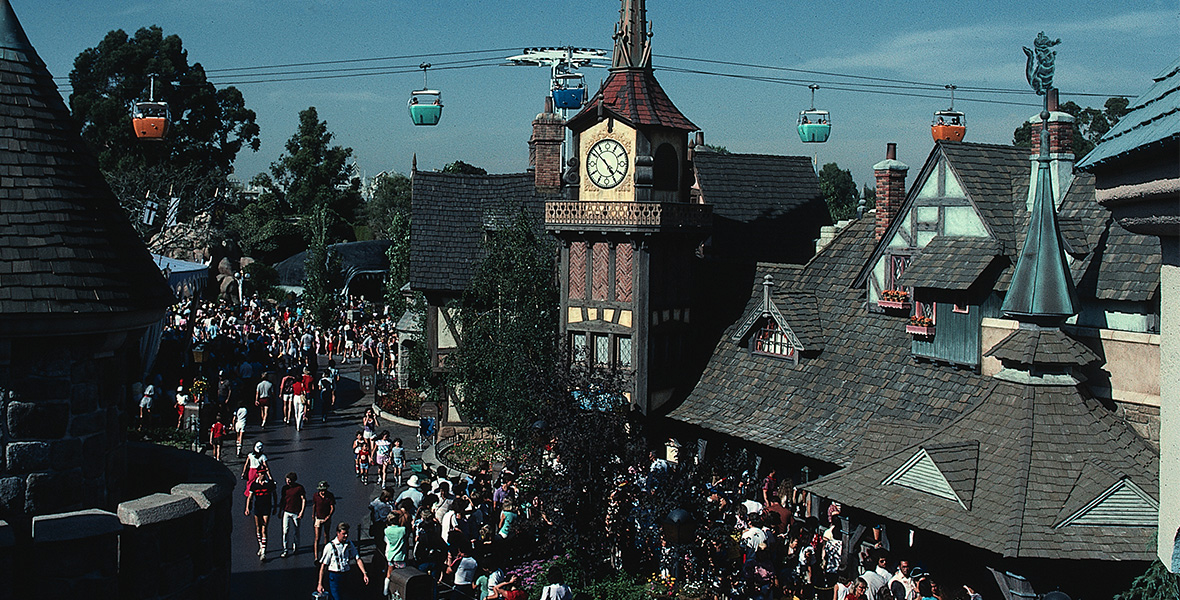 Une foule se promène dans un village à thème situé dans le New Fantasyland de Disneyland, où l’on peut admirer des gondoles colorées suspendues dans les airs et une tour horloge au centre. L’ambiance y est animée et trépidante, reflétant toute la magie qui attire les visiteurs depuis le 25 mai 1983, tandis que les rues débordantes de vie grouillent de monde.
