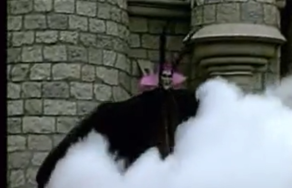 Une personne vêtue d’un costume noir et d’une coiffe rose se tient devant un mur de pierre, partiellement dissimulée par une épaisse fumée blanche — une scène qui rappelle l’ambiance spectaculaire qui règne à New Fantasyland, à Disneyland.