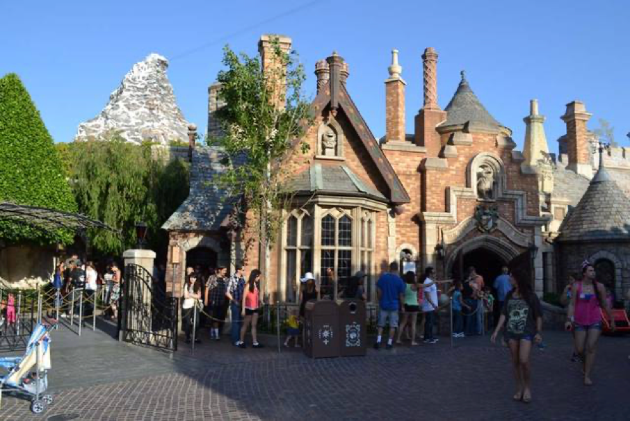 Des visiteurs se promènent et se rassemblent devant un bâtiment en briques doté de cheminées dans un parc d'attractions, avec en arrière-plan la structure emblématique du Cervin, visible sous un ciel dégagé. Cette scène animée illustre tout le charme de New Fantasyland, inauguré à Disneyland en 1983.