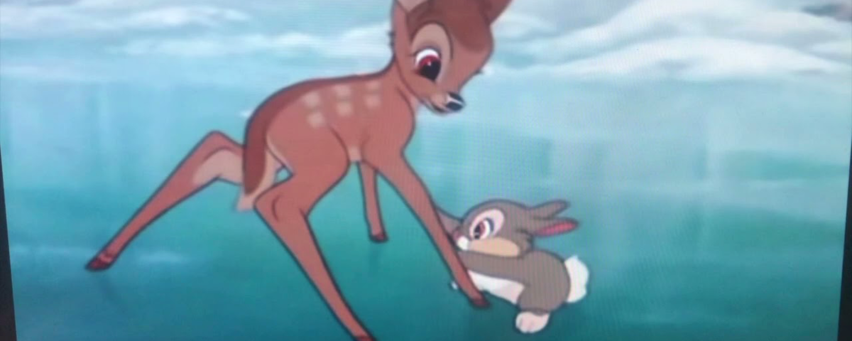 Un jeune chevreuil glisse sur la glace tandis qu’un petit lapin gris tente de le soutenir ; leur tendre complicité est un chant éternel et discret d’amour au cœur de la forêt sereine. On aperçoit de la neige et des arbres à l’arrière-plan.