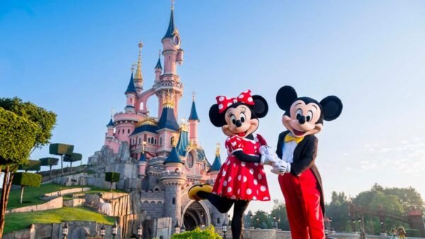 Deux personnes déguisées en Minnie Mouse et Mickey Mouse se tiennent devant le château rose de Disneyland Paris, par une journée claire et ensoleillée.