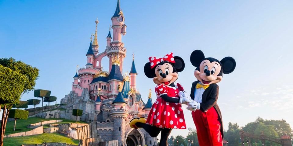 Deux personnes déguisées en Minnie Mouse et Mickey Mouse se tiennent devant le château rose de Disneyland Paris, par une journée claire et ensoleillée.