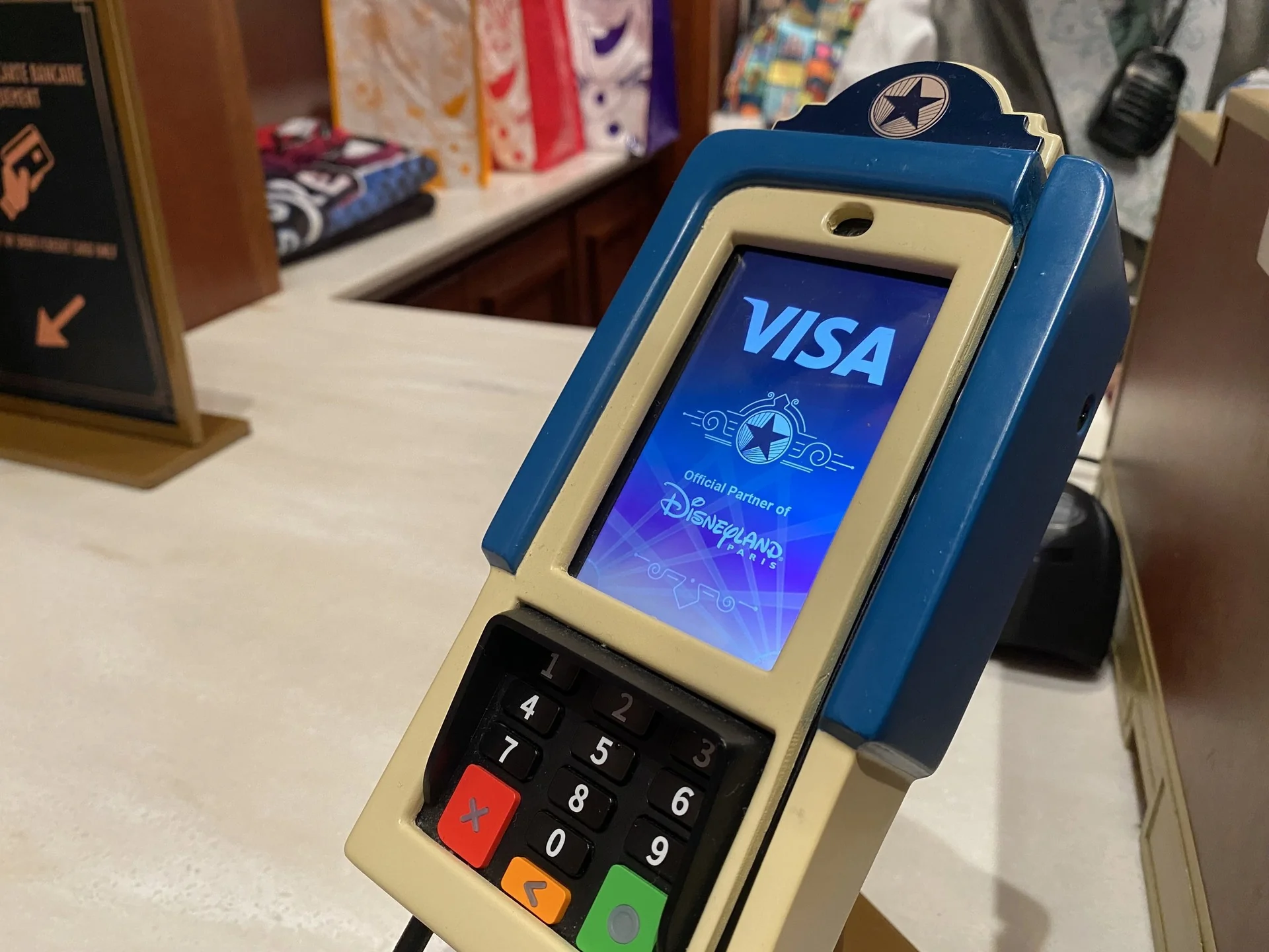 Un terminal de paiement affiche sur son écran le logo Visa et la mention « Partenaire officiel de Disneyland Paris », avec en arrière-plan des produits dérivés aux couleurs vives.