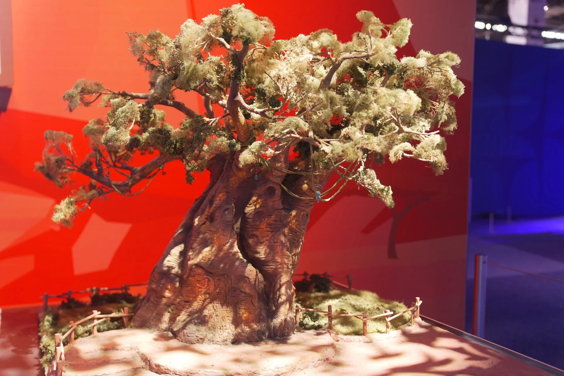 Une maquette réaliste représentant un grand arbre aux branches noueuses et au feuillage vert est exposée sur une estrade, entourée d'une petite clôture, devant un décor rouge et bleu. Cette scène immersive évoque l'univers du Roi Lion, plongeant les spectateurs dans un monde immersif qui rappelle ce film tant apprécié.