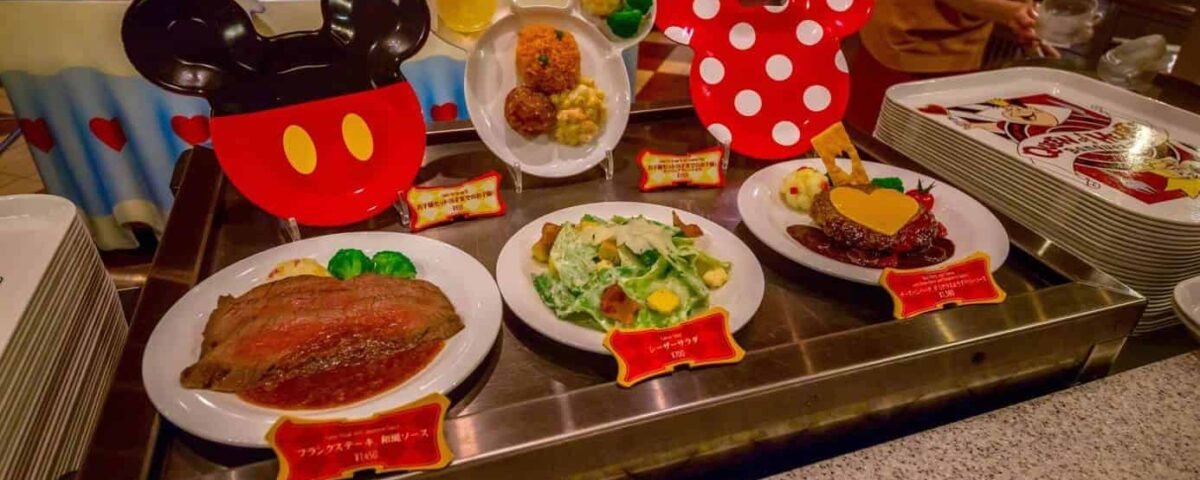 Trois plats servis à table, accompagnés de salades et de desserts, chacun présenté devant des décorations en forme d’oreilles de Mickey et Minnie, sont disposés sur un comptoir métallique dans un restaurant de style cantine — exactement le genre d’expérience culinaire ravissante que vous pourriez vivre en réservant un restaurant à Tokyo Disney.