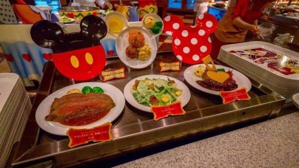 Trois plats servis à table, accompagnés de salades et de desserts, chacun présenté devant des décorations en forme d’oreilles de Mickey et Minnie, sont disposés sur un comptoir métallique dans un restaurant de style cantine — exactement le genre d’expérience culinaire ravissante que vous pourriez vivre en réservant un restaurant à Tokyo Disney.