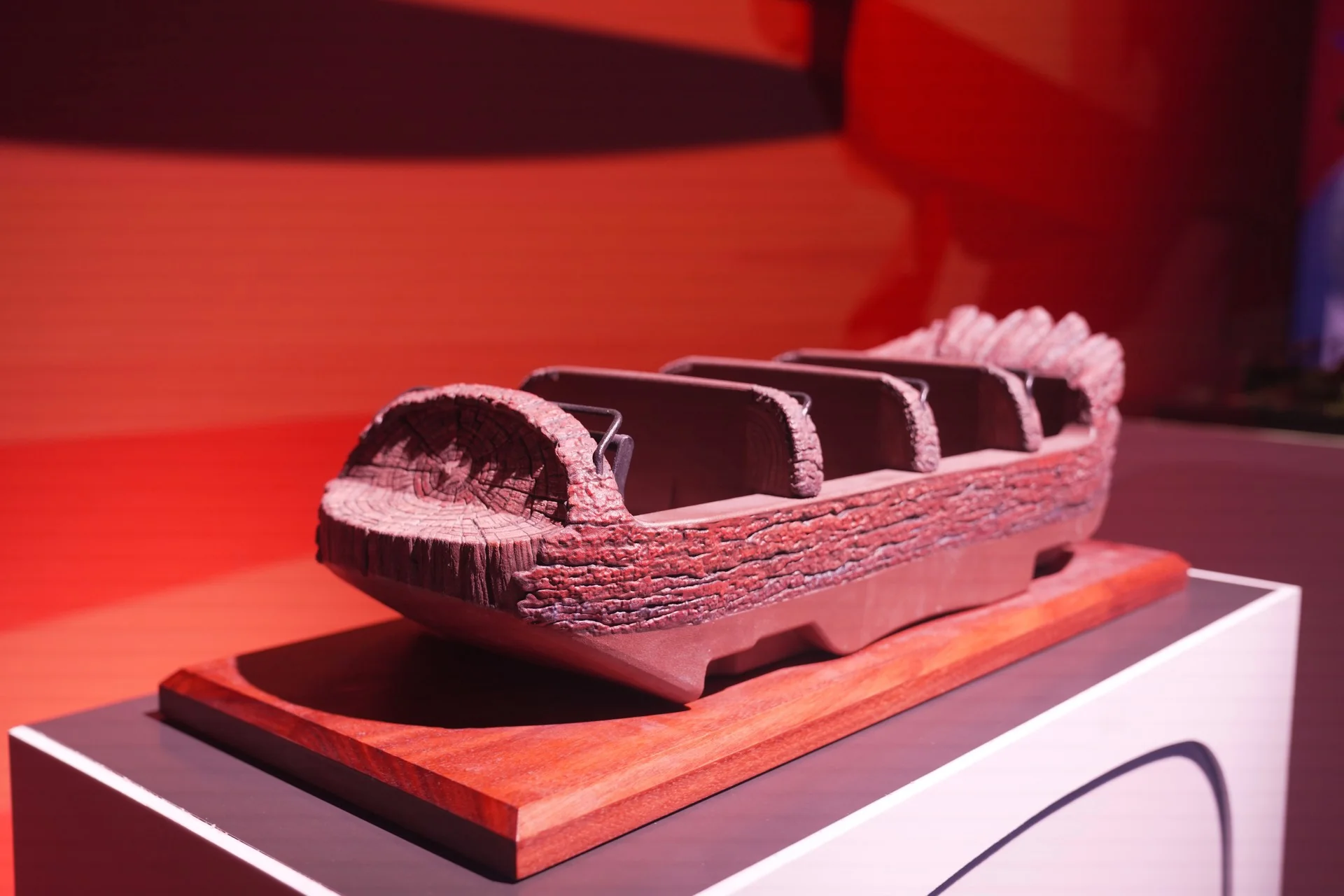 Une maquette représentant un véhicule de manège en forme de bûche, doté de quatre sièges, est exposée sur une plate-forme en bois devant un fond rouge, évoquant l'univers créatif des maquettes inspirées du « futur monde immersif ».