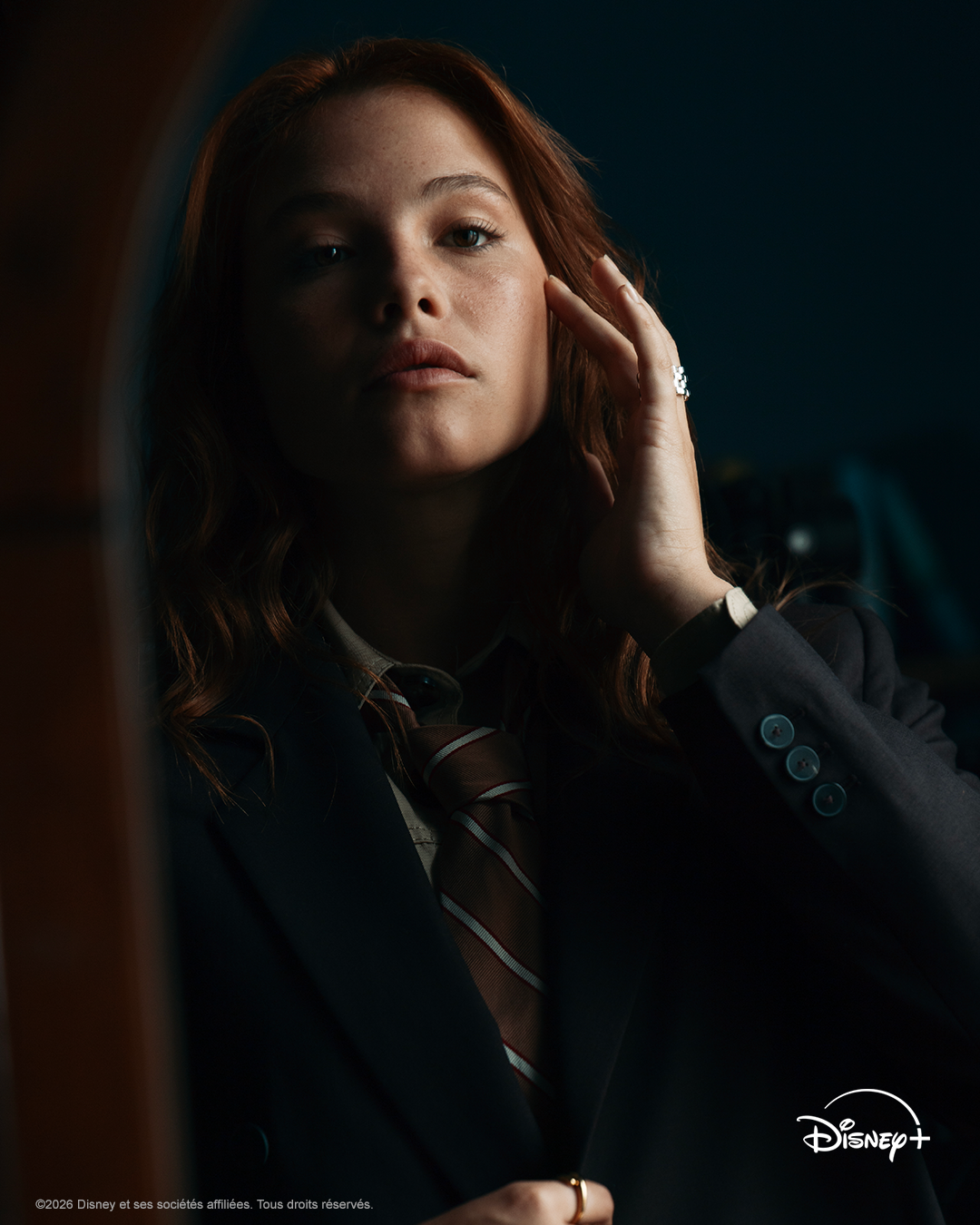 Une personne aux longs cheveux roux, vêtue d'un costume sombre et d'une cravate rayée, contemple son reflet dans un miroir, faisant écho à l'atmosphère introspective d'« Automne Pour Te Pardonner ». Le logo Disney+ apparaît dans un coin.