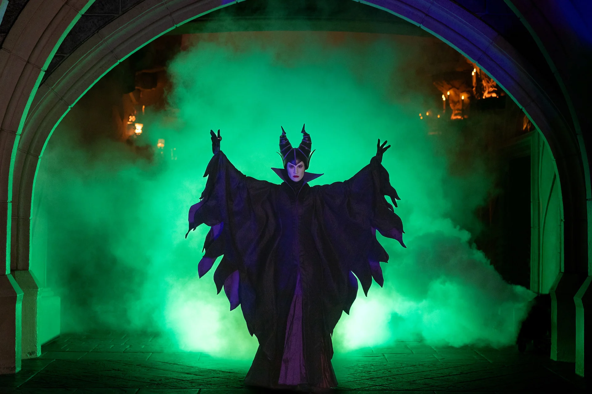 Une personne vêtue d’un costume noir spectaculaire orné de cornes se tient sous une arche, les bras levés, avec en arrière-plan une brume verte et des lumières tamisées — une image qui incarne à merveille l’esprit enchanteur du Festival d’Halloween à Disneyland Paris en 2024.
