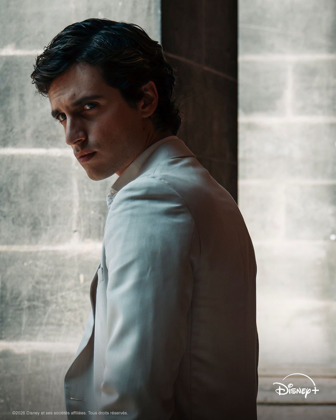 Un homme en costume blanc se tient à l'intérieur, jetant un regard par-dessus son épaule en direction de la caméra, l'air grave. Le logo Disney+ dans le coin laisse entrevoir une nouvelle sortie, peut-être inspirée par l'intrigue de « Roman Phénomène » ou par la tension dramatique d'« Un Automne Pour Te Pardonner ».